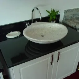 Czarny blat kamienny z owalną, jasną umywalką z kamienia, chromowaną baterią i małą doniczką z zieloną rośliną, zamontowany na białej szafce z frezowanymi frontami.