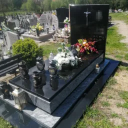 Czarny, błyszczący pomnik z kwiatami i zniczami na cmentarzu w słoneczny dzień. Widoczny metalowy krzyż i wyryte dane zmarłych. Uwagę przyciągają lampiony w stylu retro.