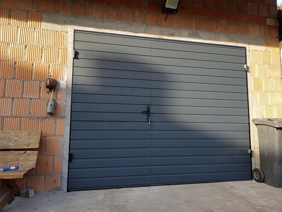 Zamknięta, segmentowa brama garażowa w kolorze grafitowym, zamontowana w surowym murze z cegły kratówki, z widocznym oświetleniem zewnętrznym i fragmentem kosza na śmieci z boku.