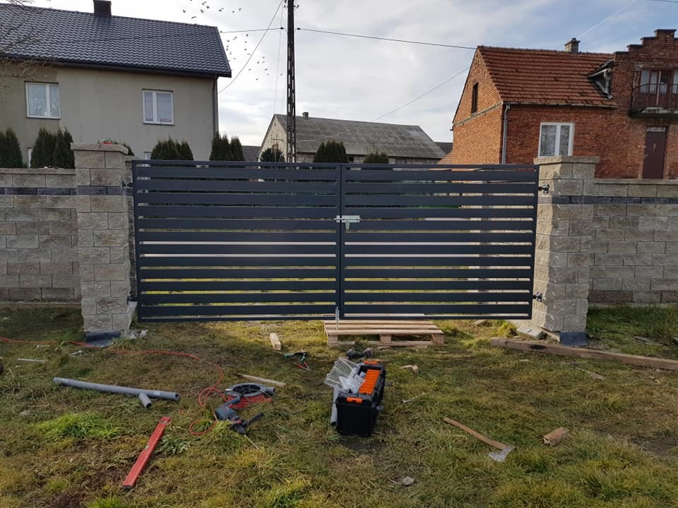 Nowoczesna brama wjazdowa z poziomych metalowych profili, osadzona na murowanych słupkach z szarej kostki, z widocznymi narzędziami i elementami montażowymi na trawie.