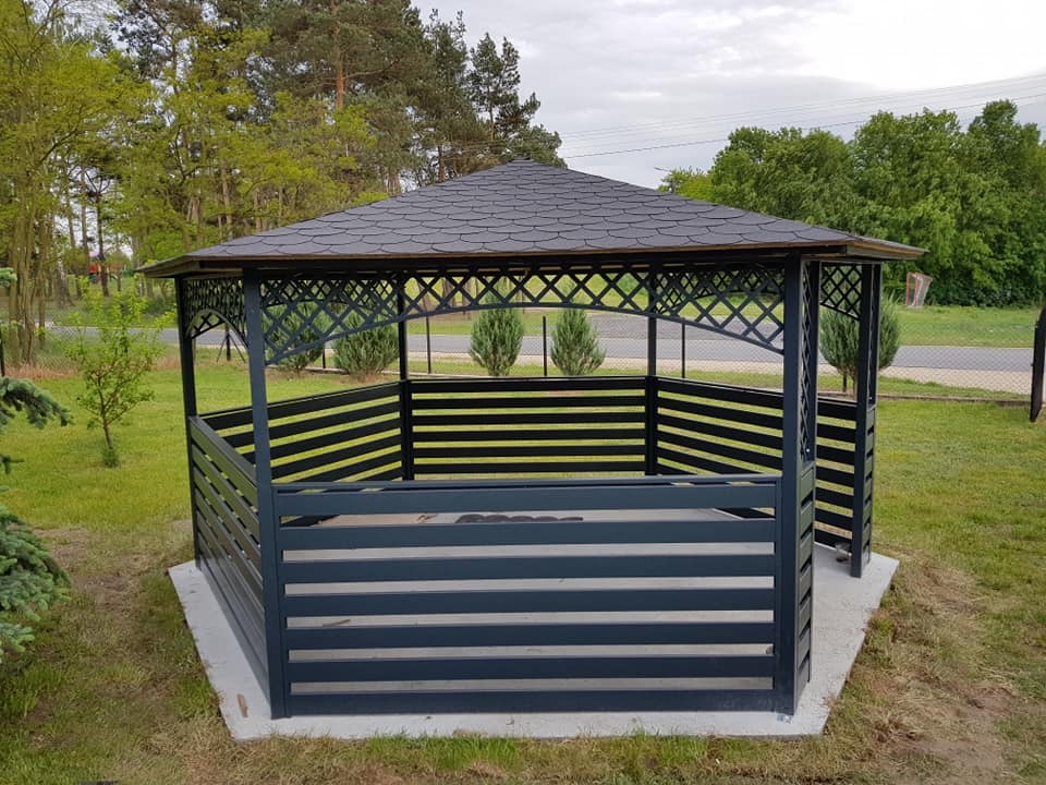 Altana ogrodowa z poziomymi panelami, czarna konstrukcja z ażurową górą, szary dach z gontu bitumicznego, otoczona zielenią.