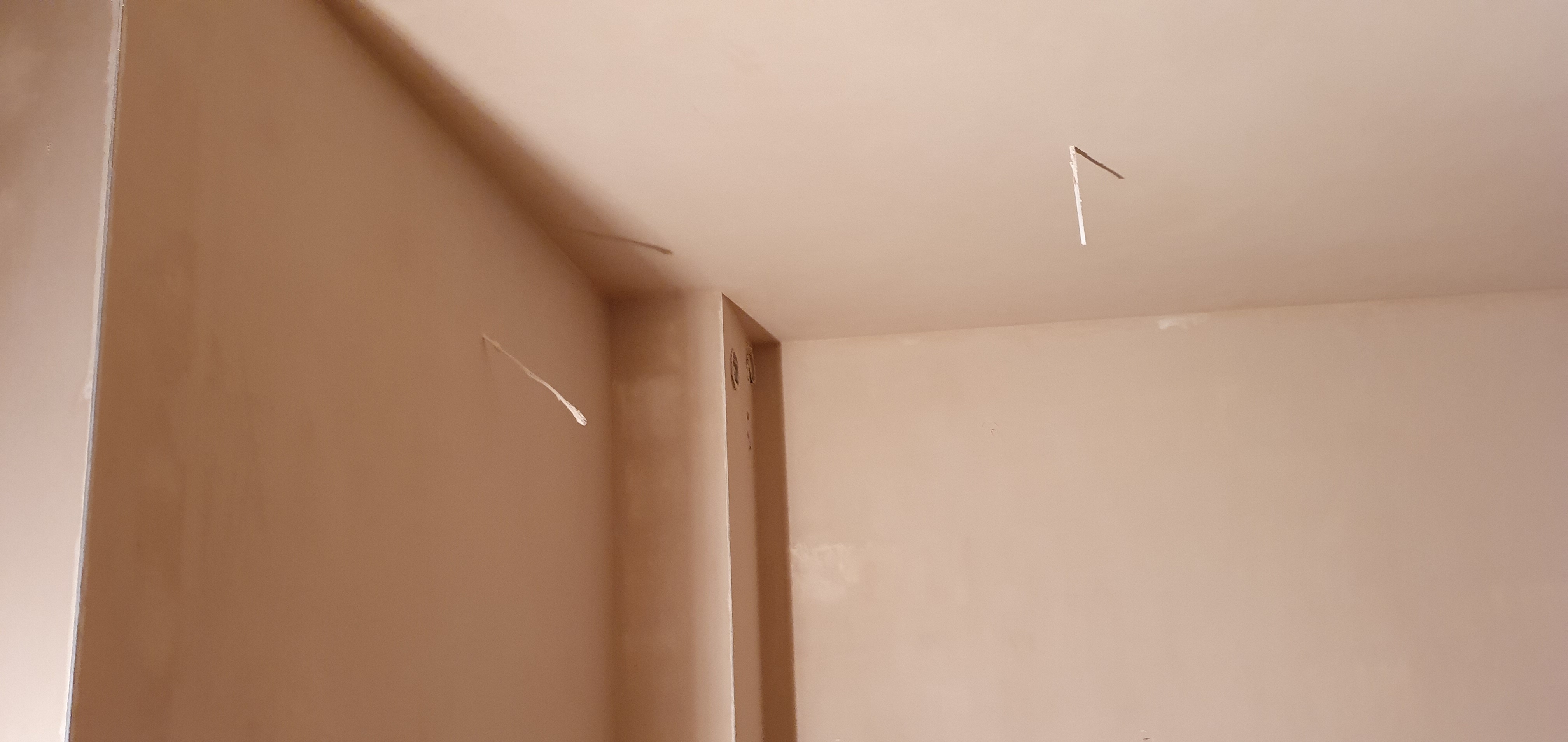 Świeżo nałożona, gładka, beżowa powłoka na ścianach i suficie w rogu pokoju, z wystającymi przewodami elektrycznymi.