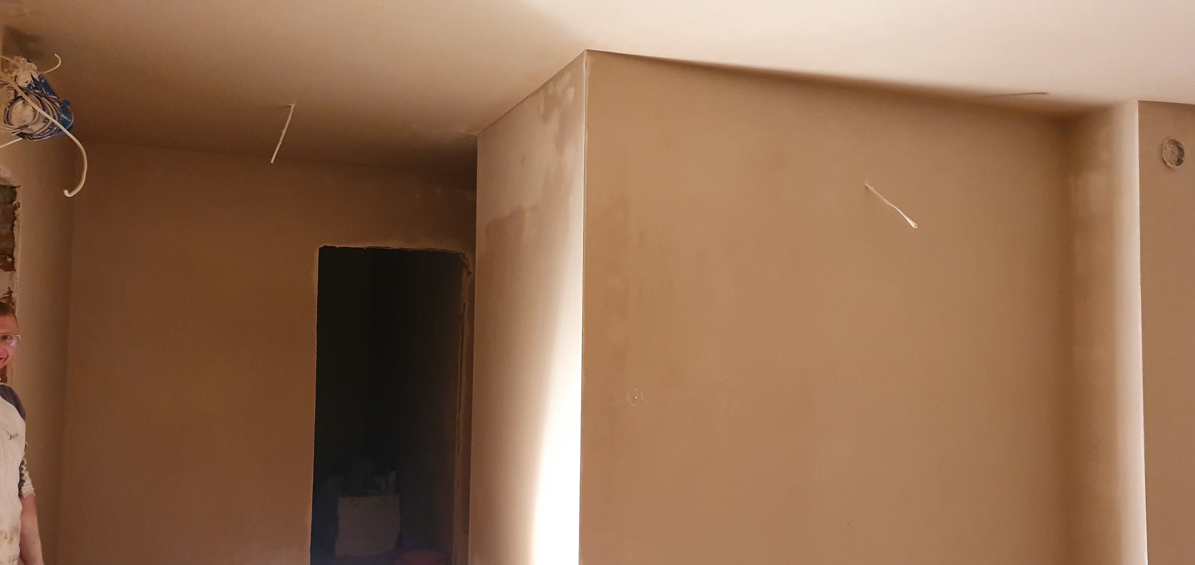 Świeżo otynkowane wnętrze z widocznymi przewodami elektrycznymi na suficie i fragmentem osoby w roboczym ubraniu po lewej stronie kadru.