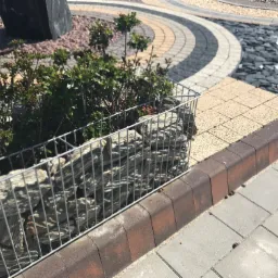 Gabion z kamieniami i roślinami, obok kostki brukowej w różnych kolorach i wzorach, na zewnątrz.