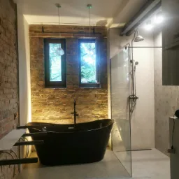 Nowoczesna łazienka z czarną wanną wolnostojącą na tle ściany z czerwonej cegły i przeszklonym prysznicem typu walk-in.