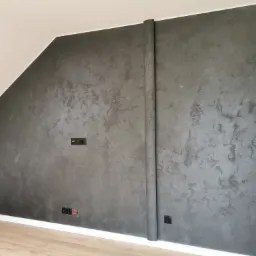 Ściana w pokoju na poddaszu pokryta dekoracyjną szarą farbą z efektem betonu, widoczne gniazdka elektryczne i wentylacja, listwa przypodłogowa, jasna podłoga.