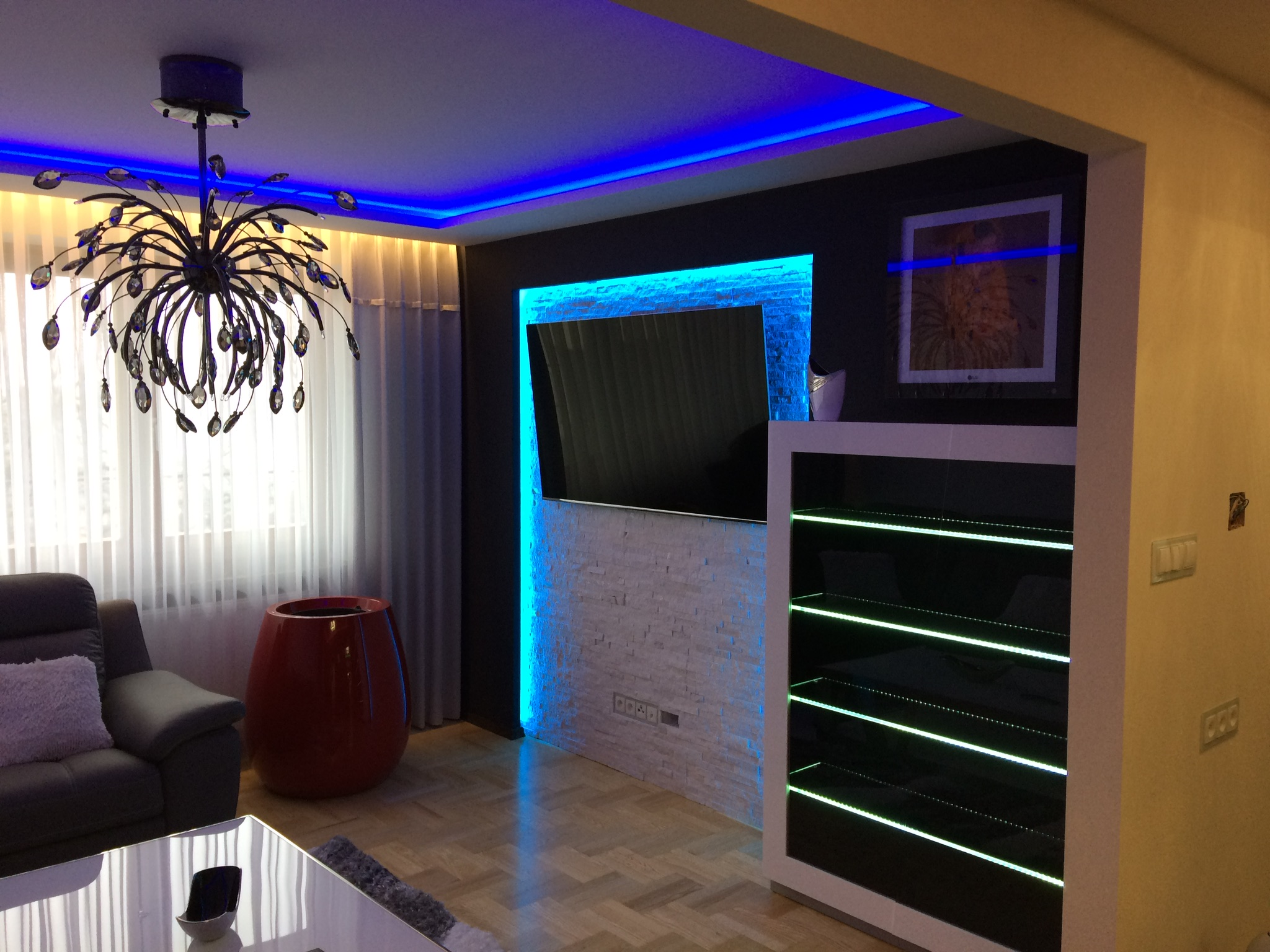 Nowoczesny salon z podświetlanym telewizorem na ścianie z imitacji kamienia, designerski żyrandol i szafka z podświetlanymi półkami, tworzące nastrojowe oświetlenie wnętrza.