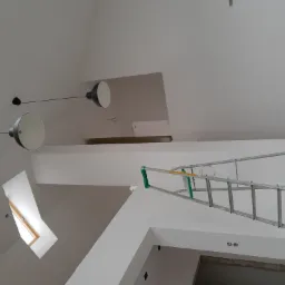 Skośne poddasze w trakcie prac wykończeniowych, widoczne okno dachowe, aluminiowa drabina oparta o ścianę, surowe ściany przygotowane do malowania i wiszące lampy.