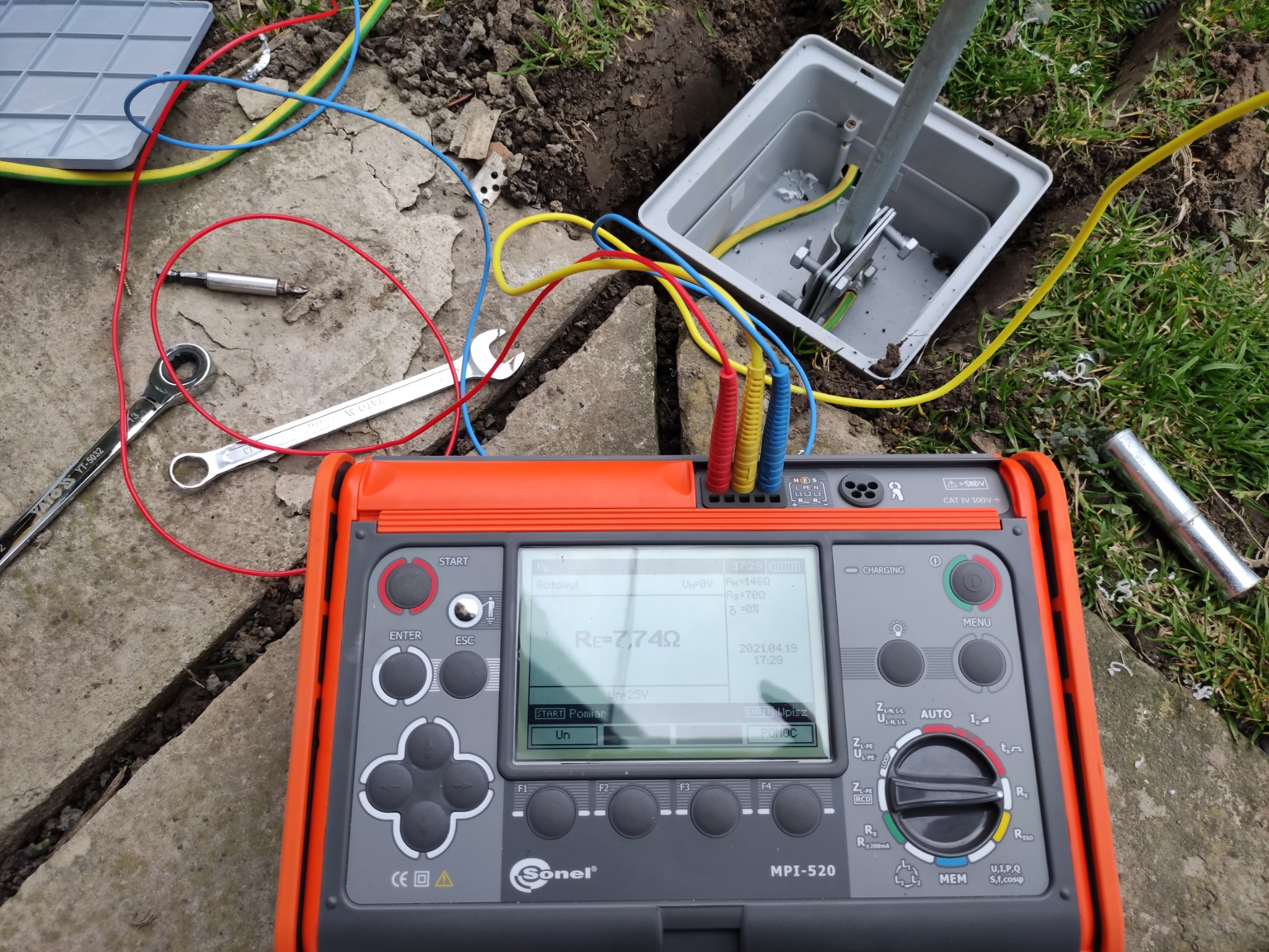 Miernik Sonel MPI-520 podczas pomiaru rezystancji uziomu, widoczne kable pomiarowe, skrzynka przyłączeniowa uziomu, narzędzia i fragment trawnika.