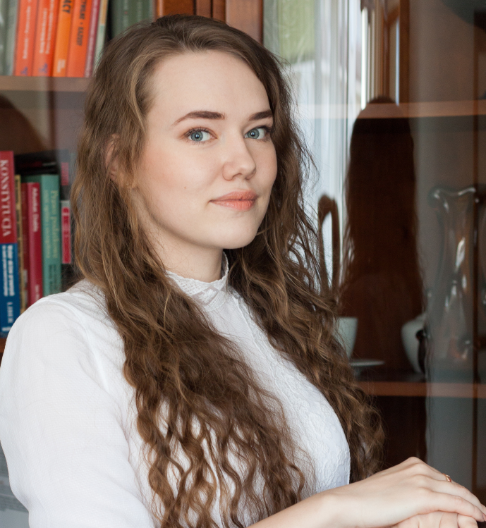 Portret młodej kobiety z długimi kręconymi włosami, ubranej w białą bluzkę, na tle przeszklonej biblioteczki z książkami.