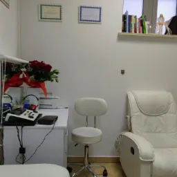 Gabinet z białymi meblami, krzesłem na kółkach, fotelem masującym i sprzętem diagnostycznym na biurku, ozdobiony bukietem czerwonych róż i certyfikatami na ścianie.