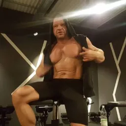 Masywny mężczyzna z odsłoniętym torsem w czarnej bluzie z kapturem, siedzący na maszynie do ćwiczeń w siłowni, prezentujący napięte mięśnie brzucha i klatki piersiowej.