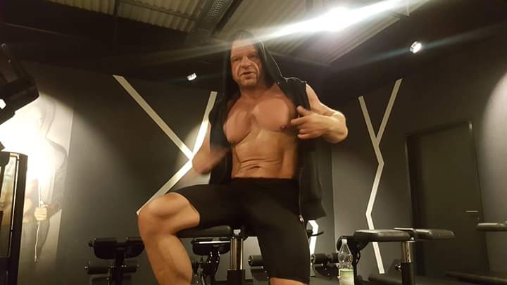 Masywny mężczyzna z odsłoniętym torsem w czarnej bluzie z kapturem, siedzący na maszynie do ćwiczeń w siłowni, prezentujący napięte mięśnie brzucha i klatki piersiowej.