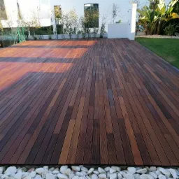 Nowoczesny taras z egzotycznego drewna, otoczony białymi kamieniami i zielenią ogrodu, w słoneczny dzień.