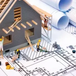 Miniaturowy model domu w trakcie budowy, z figurkami robotników, betoniarką i taczką, umieszczony na planach architektonicznych.