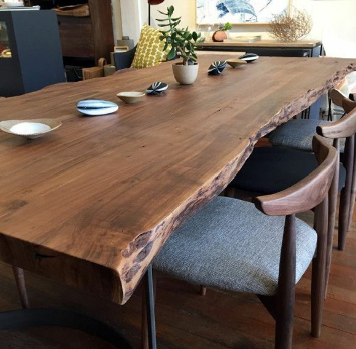 Stół z litego drewna o nieregularnej krawędzi, wykończony na wysoki połysk, na metalowych nogach, otoczony krzesłami z drewnianymi oparciami i szarą tapicerką, udekorowany miseczkami i rośliną...