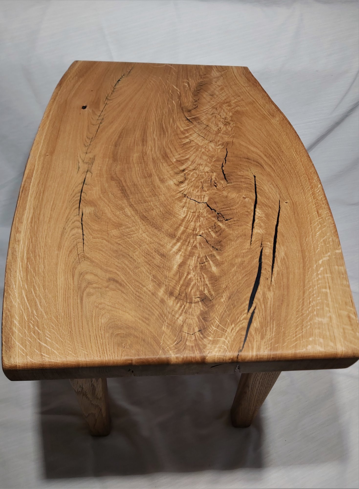 Niski, ręcznie robiony stołek z blatem z litego drewna dębowego z widocznymi pęknięciami wypełnionymi czarną żywicą, na tle białej tkaniny.
