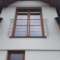 Zewnętrzna, minimalistyczna barierka okienna ze stali nierdzewnej, zamontowana na oknie poddaszowym w budynku o jasnej elewacji.