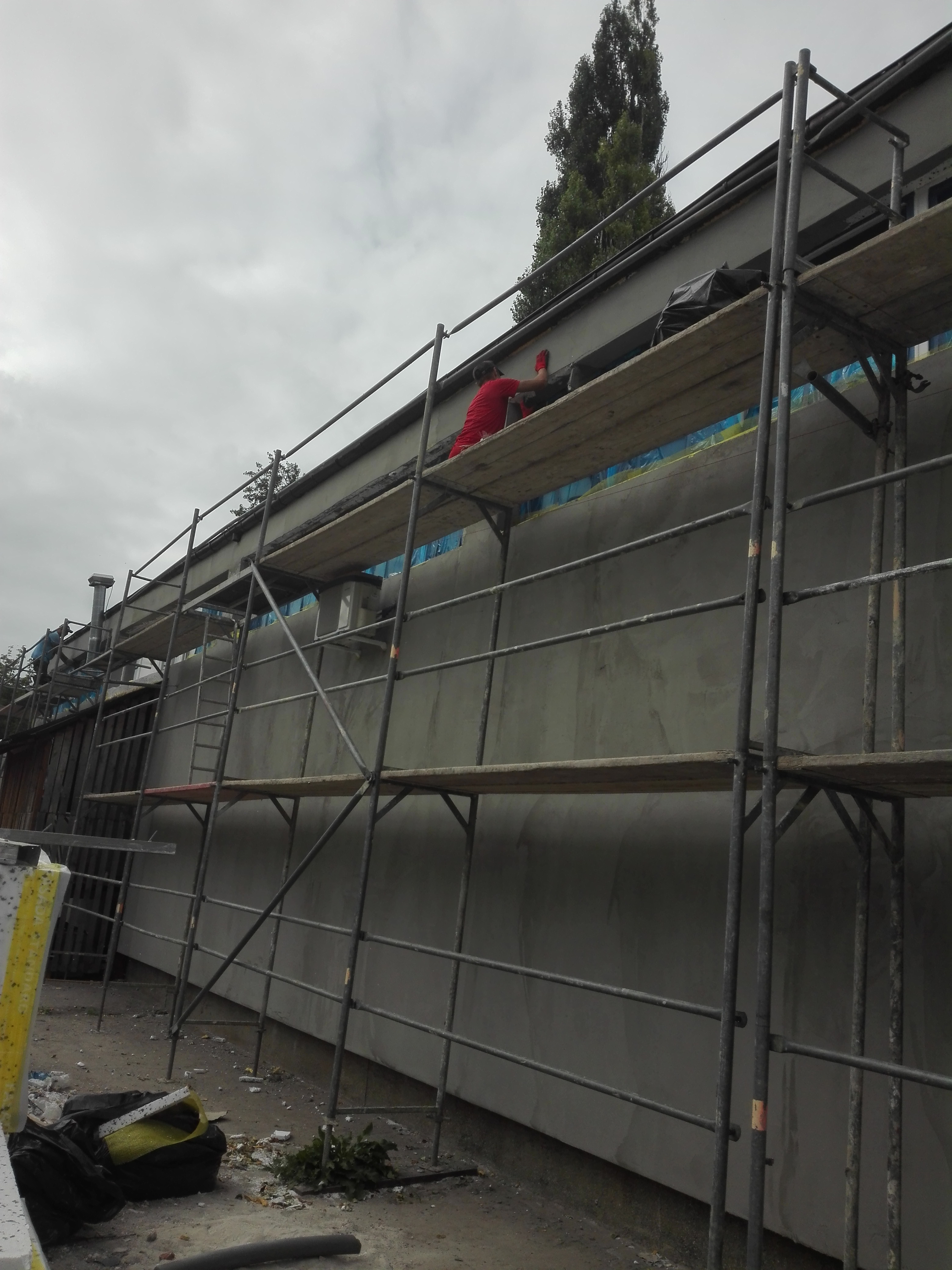 Pracownik w czerwonym kombinezonie na rusztowaniu, wygładza szarą, surową elewację budynku. W tle widoczne drzewo i pochmurne niebo.