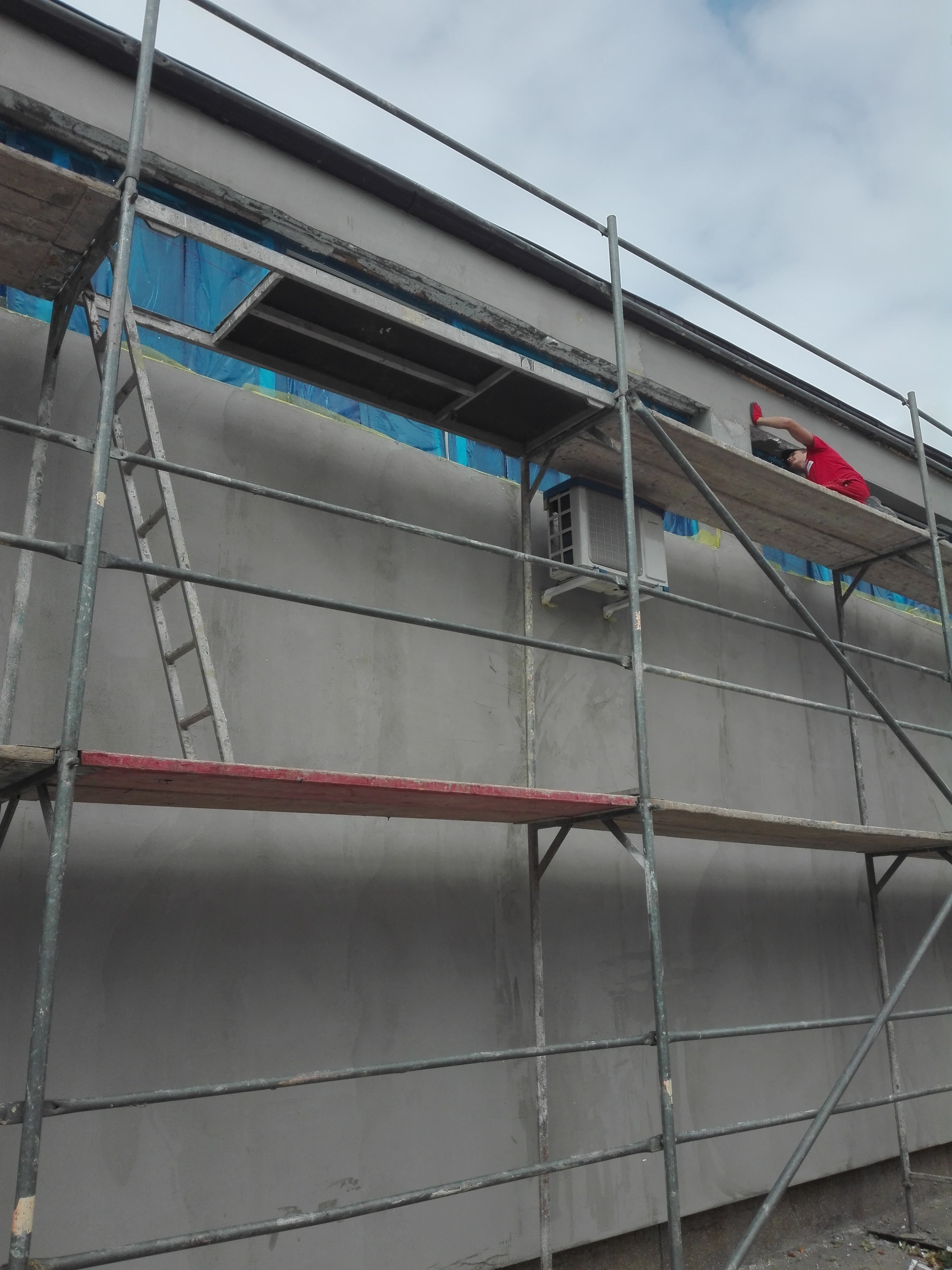 Pracownik w czerwonej koszulce na rusztowaniu, wyrównuje szarą elewację budynku, okna zabezpieczone folią, widoczny klimatyzator.