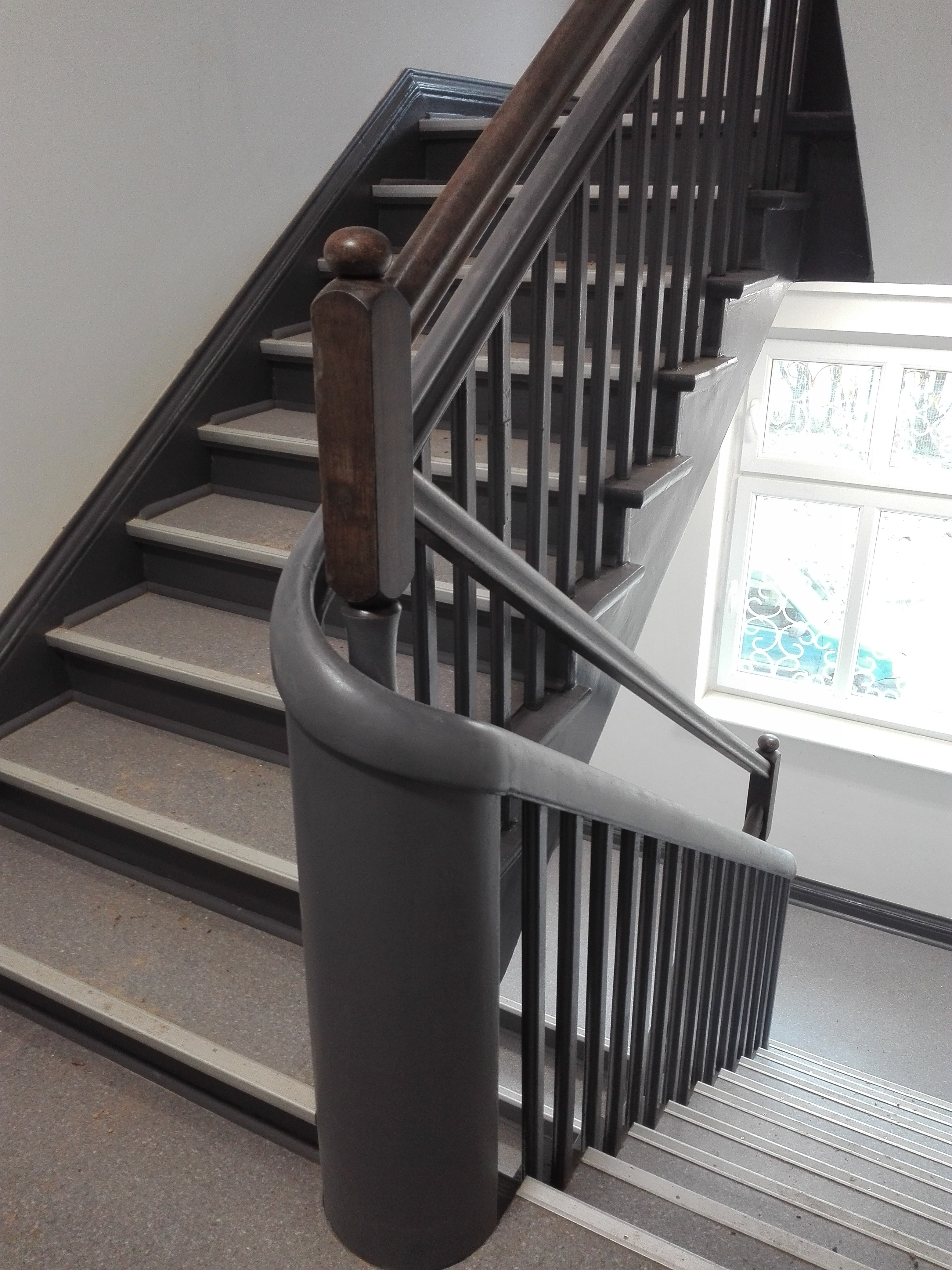 Klatka schodowa z drewnianą balustradą pomalowaną na ciemny kolor, widok z góry na schody z szarymi stopniami i białymi listwami, okno z ozdobnymi szprosami w tle.