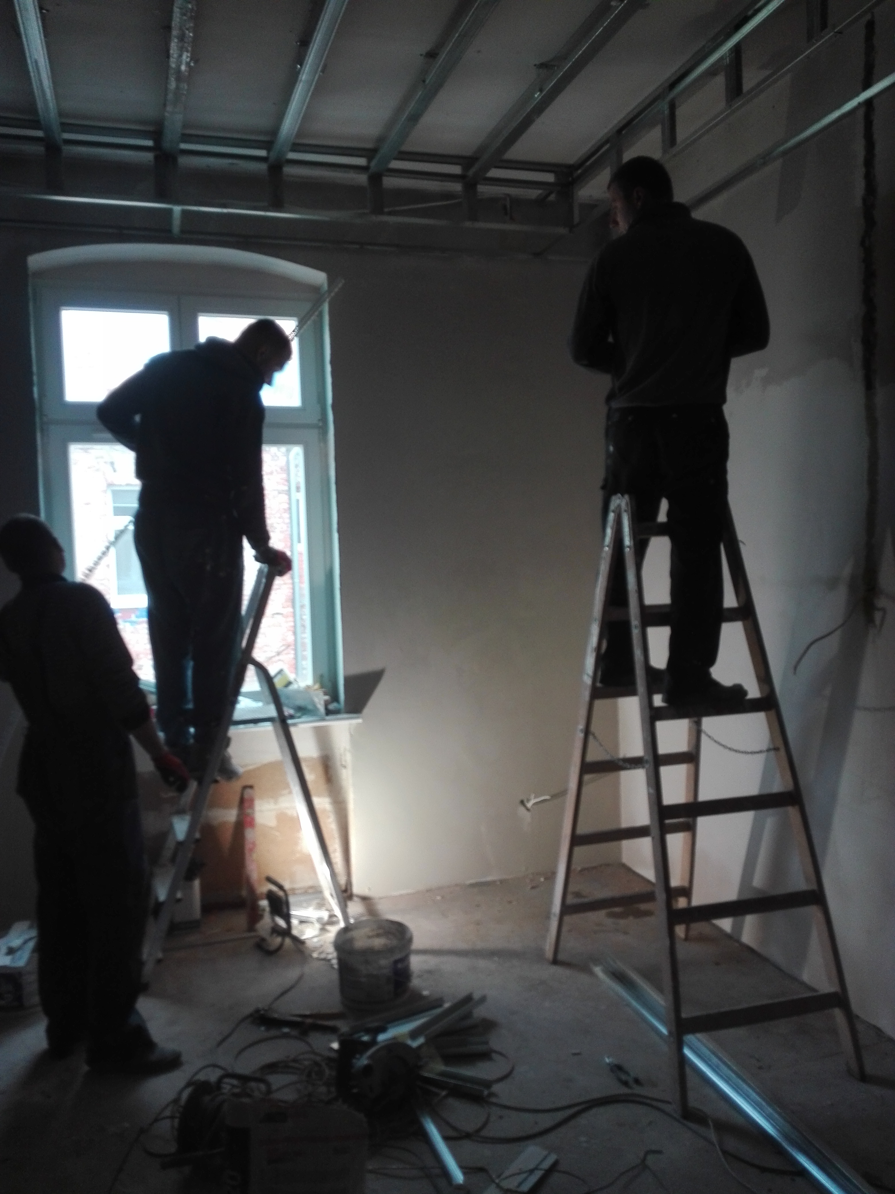 Montaż sufitu podwieszanego: trzech pracowników na drabinach instaluje metalową konstrukcję sufitu podwieszanego w pokoju z oknem, widoczne narzędzia i materiały budowlane na podłodze.