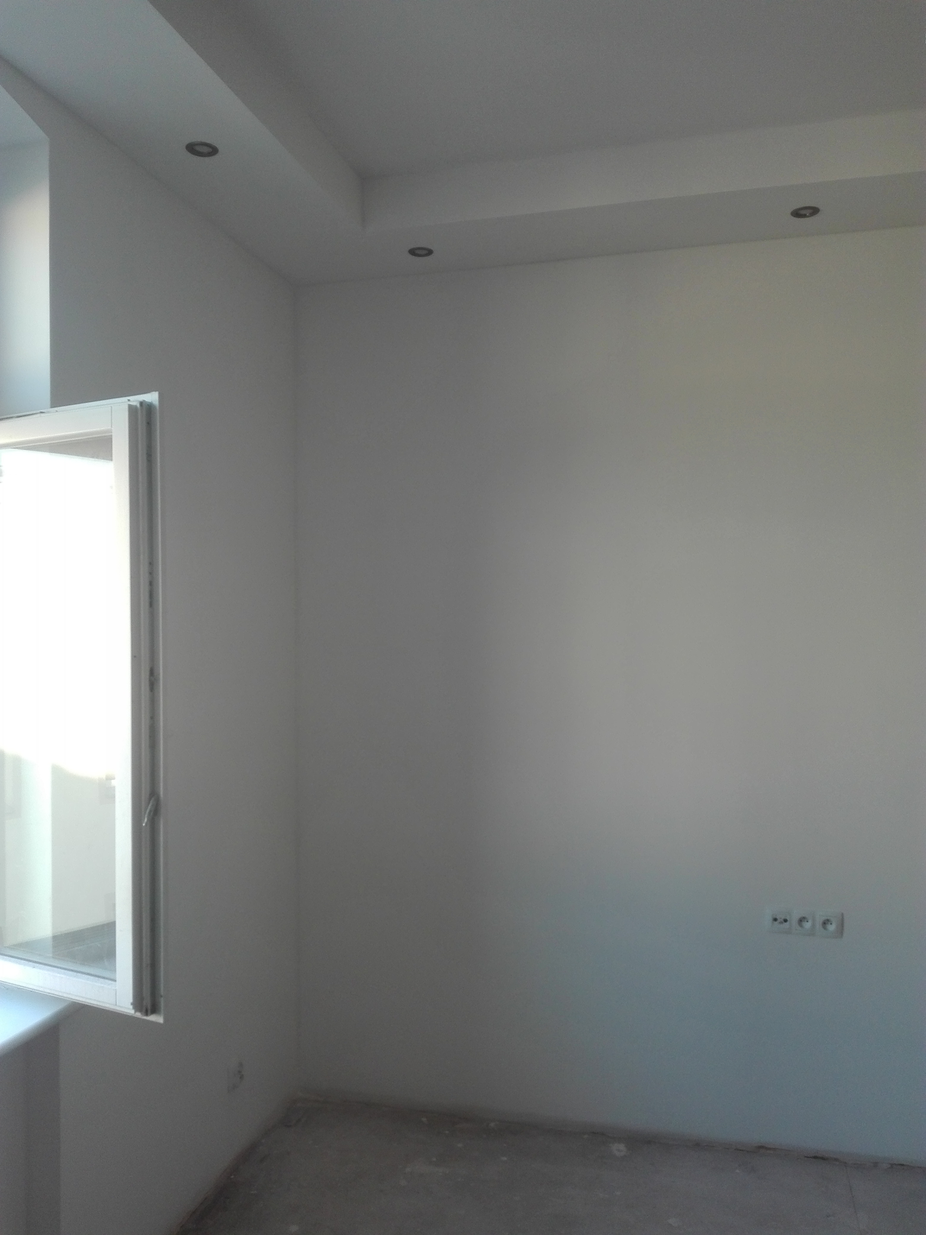 Świeżo pomalowane na biało wnętrze z oknem po lewej stronie i trzema gniazdkami elektrycznymi na ścianie po prawej, widoczne oświetlenie sufitowe.