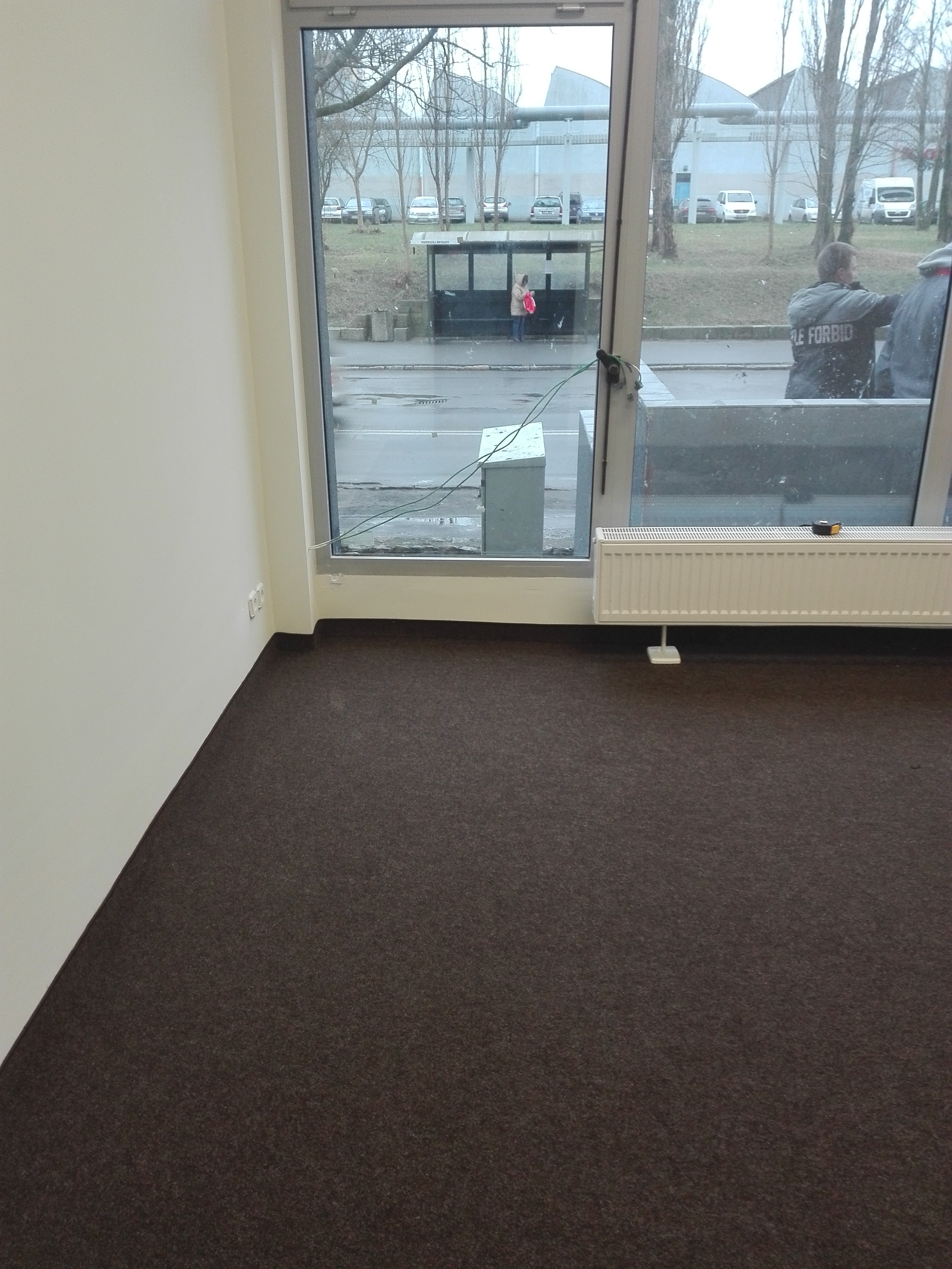 Wnętrze pokoju w Szczecinie z brązową wykładziną dywanową, grzejnikiem pod oknem i widokiem na przystanek autobusowy.