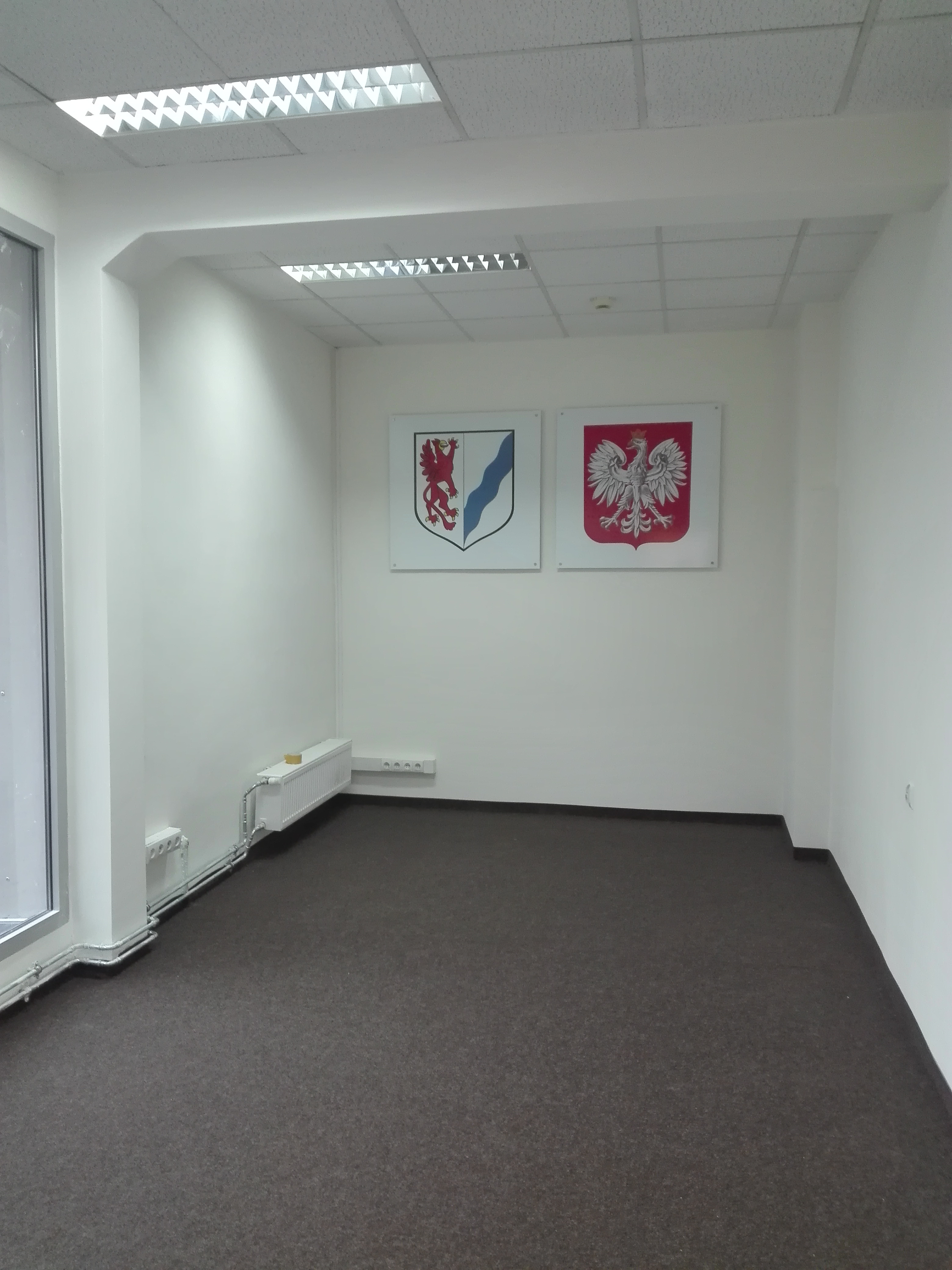 Puste, jasne biuro z wykładziną dywanową i herbami na ścianie, oświetlone kasetonowymi lampami sufitowymi.