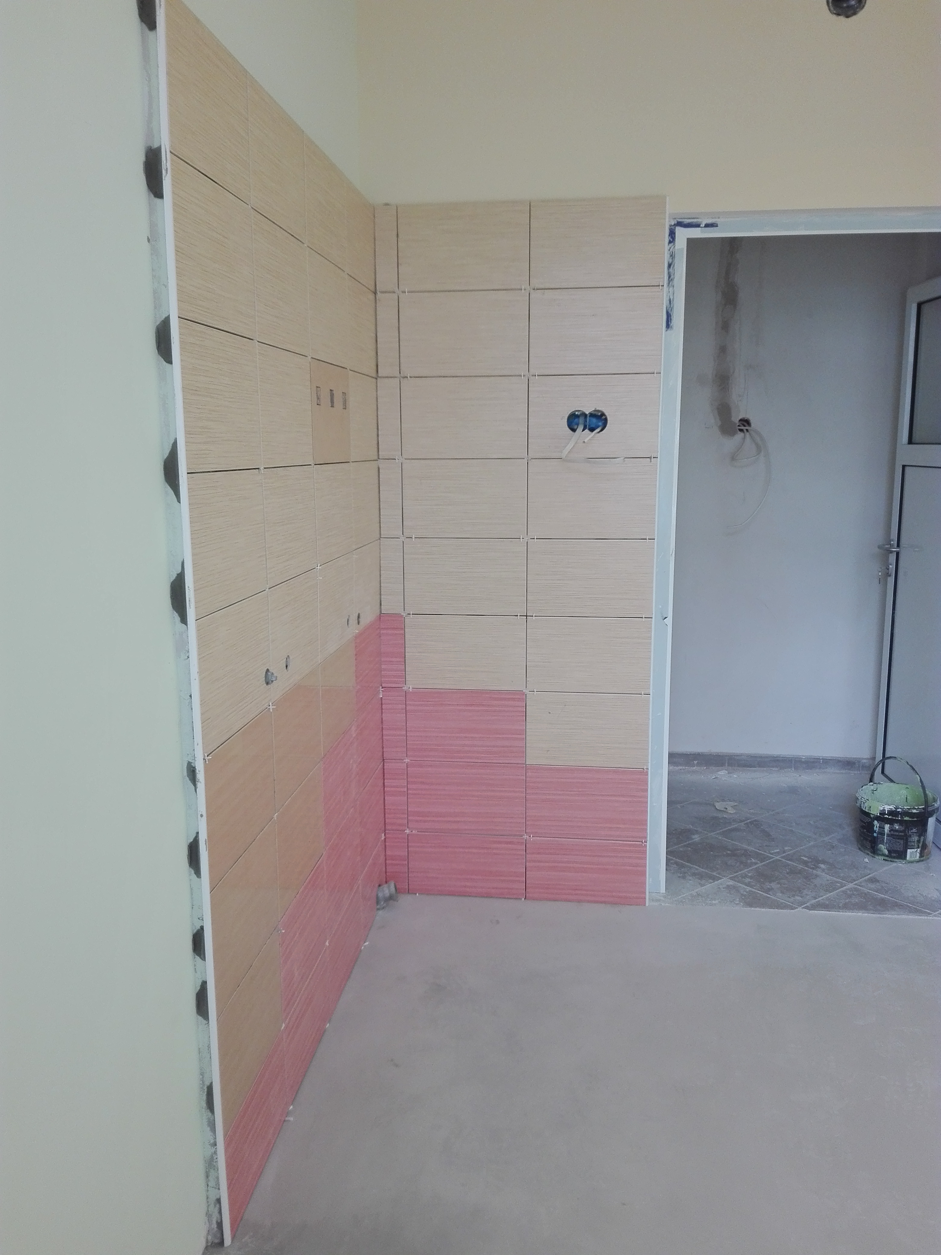 Wnętrze w trakcie układania płytek ściennych w dwóch odcieniach: beżowym i różowym, widoczne puszki instalacyjne z przewodami, fragment podłogi z szarym podkładem oraz pomieszczenie obok...