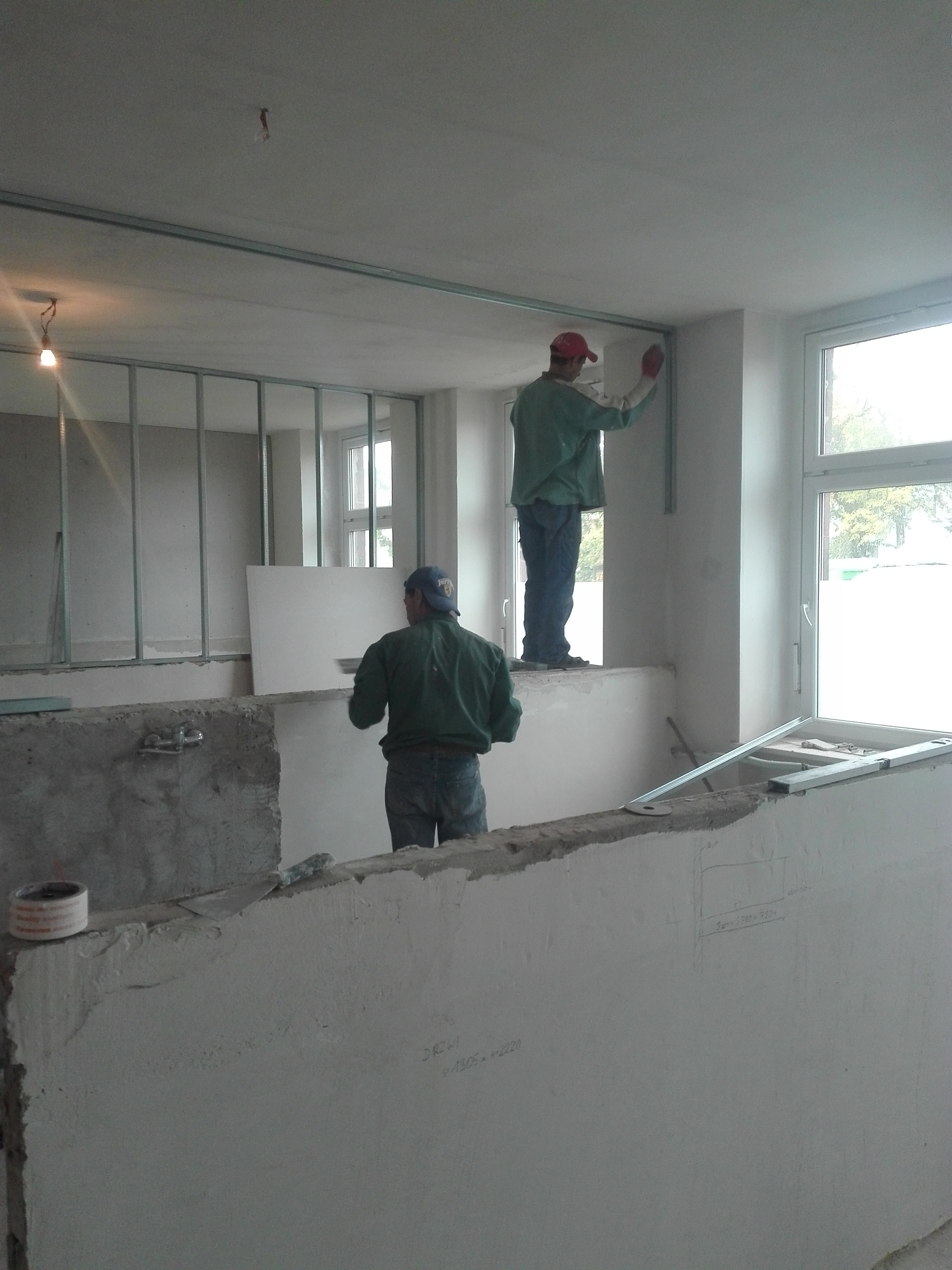 Dwóch mężczyzn w trakcie montażu metalowej konstrukcji pod płyty gipsowo-kartonowe wewnątrz pomieszczenia, widoczne elementy instalacji elektrycznej i surowe ściany.