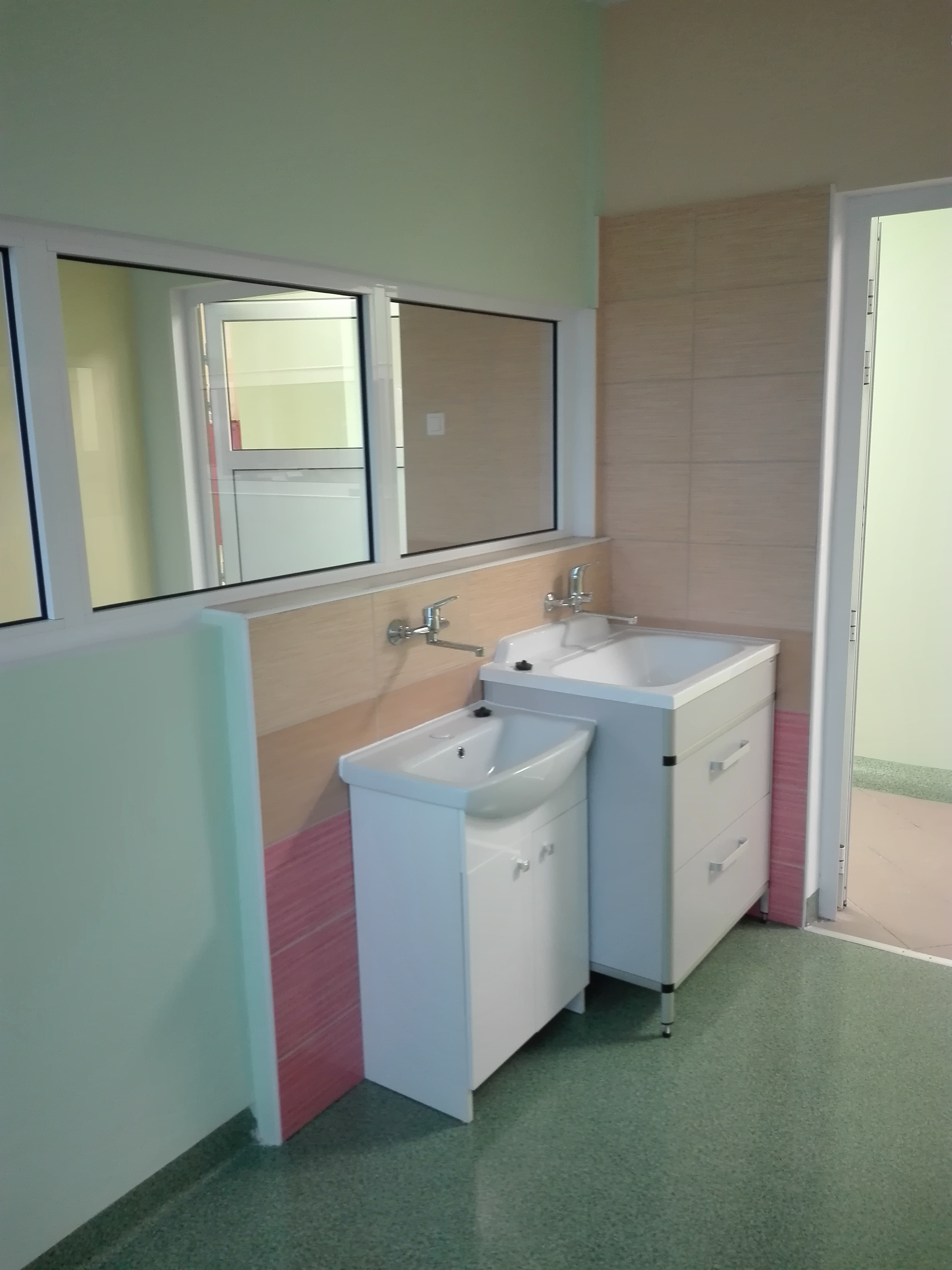 Dwie białe umywalki z szafkami, jedna z szufladami, zamontowane w łazience z beżowymi płytkami i zieloną podłogą, widok z boku, lustro nad umywalkami odbija wnętrze pomieszczenia.