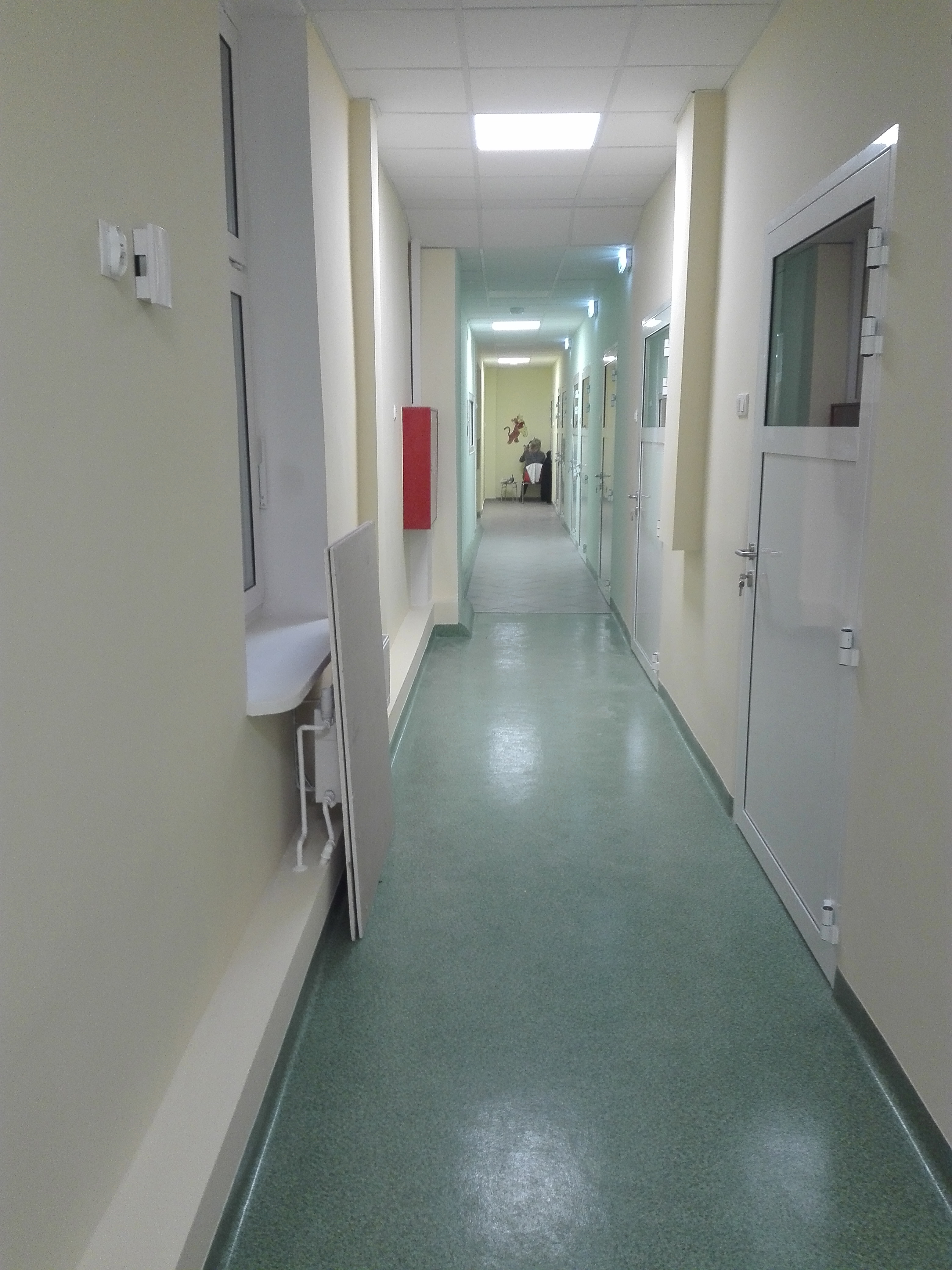 Długi, oświetlony korytarz z zieloną podłogą w budynku użyteczności publicznej, z białymi ścianami i drzwiami po obu stronach, widoczny element instalacji sanitarnej z umywalką przy ścianie po...