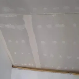 Skośny sufit poddasza w trakcie wykańczania, widoczne łączenia płyt zaszpachlowane, ale przed ostatecznym szlifowaniem, z widocznymi śladami po wkrętach.