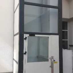 Zewnętrzna winda dla osób niepełnosprawnych, z przeszklonym szybem i białymi panelami, zamontowana przy budynku, z widocznym panelem sterowania i naklejką informacyjną.