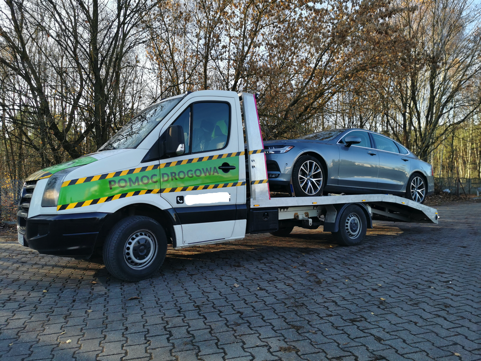Biała ciężarówka pomocy drogowej z zielonymi i żółtymi pasami transportuje srebrny samochód osobowy na platformie. W tle drzewa bez liści i kostka brukowa.