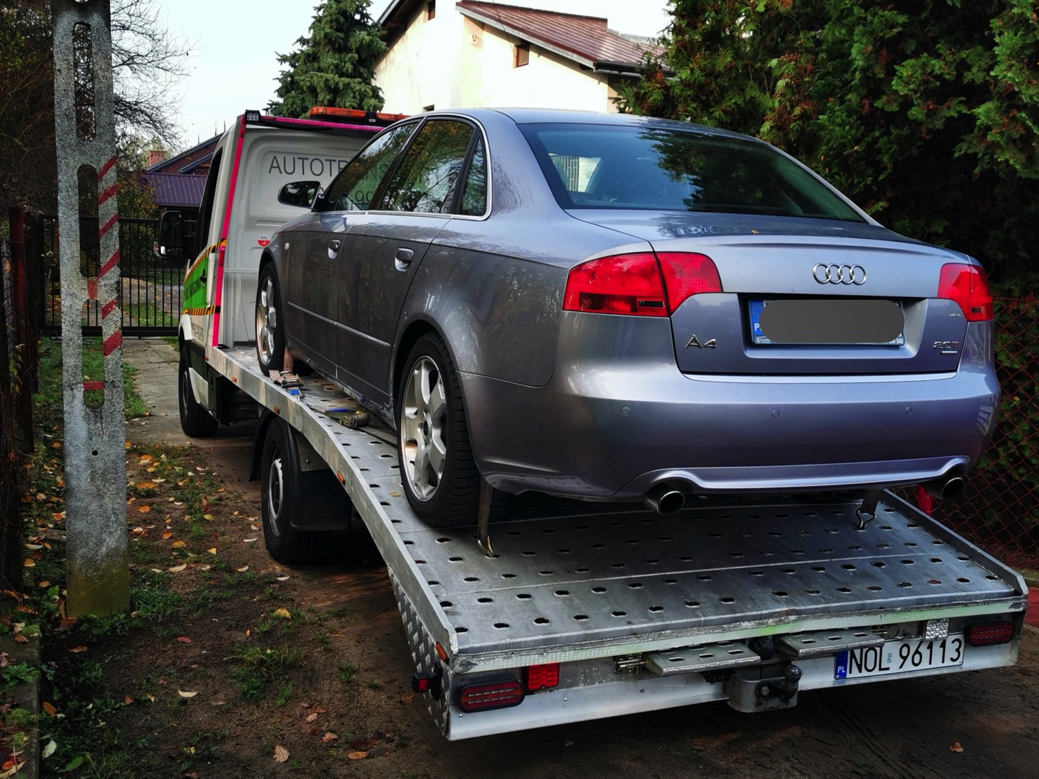 Szary samochód Audi A4 2.0T zabezpieczony na srebrnej platformie lawety z widocznym numerem rejestracyjnym, podczas transportu na tle zieleni i zabudowań.
