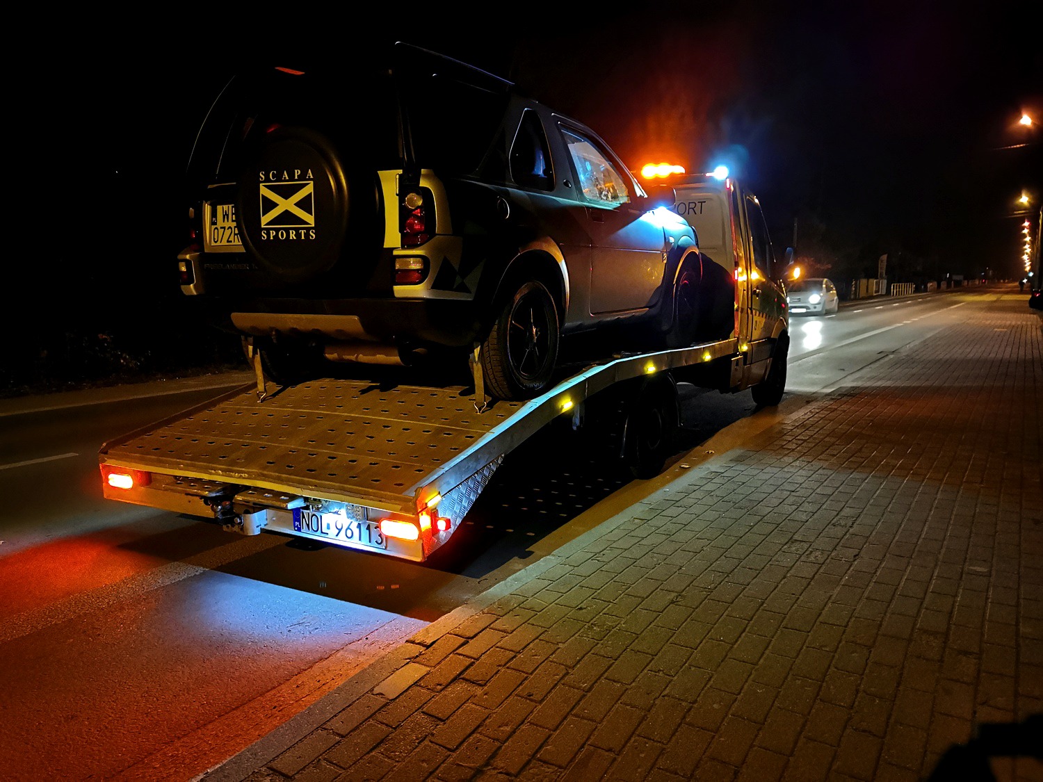 Ciemnozielony SUV marki Land Rover na platformie pojazdu pomocy drogowej, widok z tyłu, nocne ujęcie z włączonymi światłami ostrzegawczymi, tablica rejestracyjna NOL 96113.