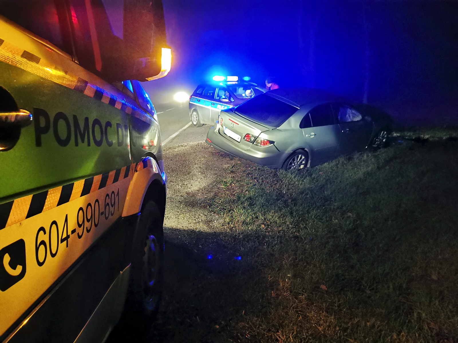 Uszkodzony, srebrny samochód po wypadku znajduje się w rowie, w tle radiowóz policyjny z włączonymi światłami i pojazd pomocy drogowej z napisem 'POMOC DROGOWA' i numerem telefonu, nocne oświetlenie.