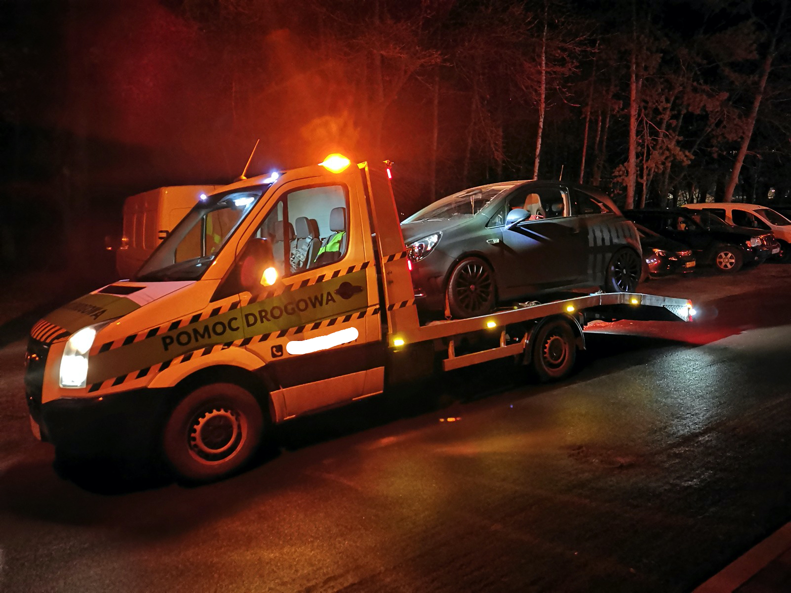Samochód osobowy w kolorze grafitowym transportowany na platformie pojazdu pomocy drogowej z napisem 'POMOC DROGOWA' w nocnych warunkach, z włączonymi światłami ostrzegawczymi. W tle widoczne...