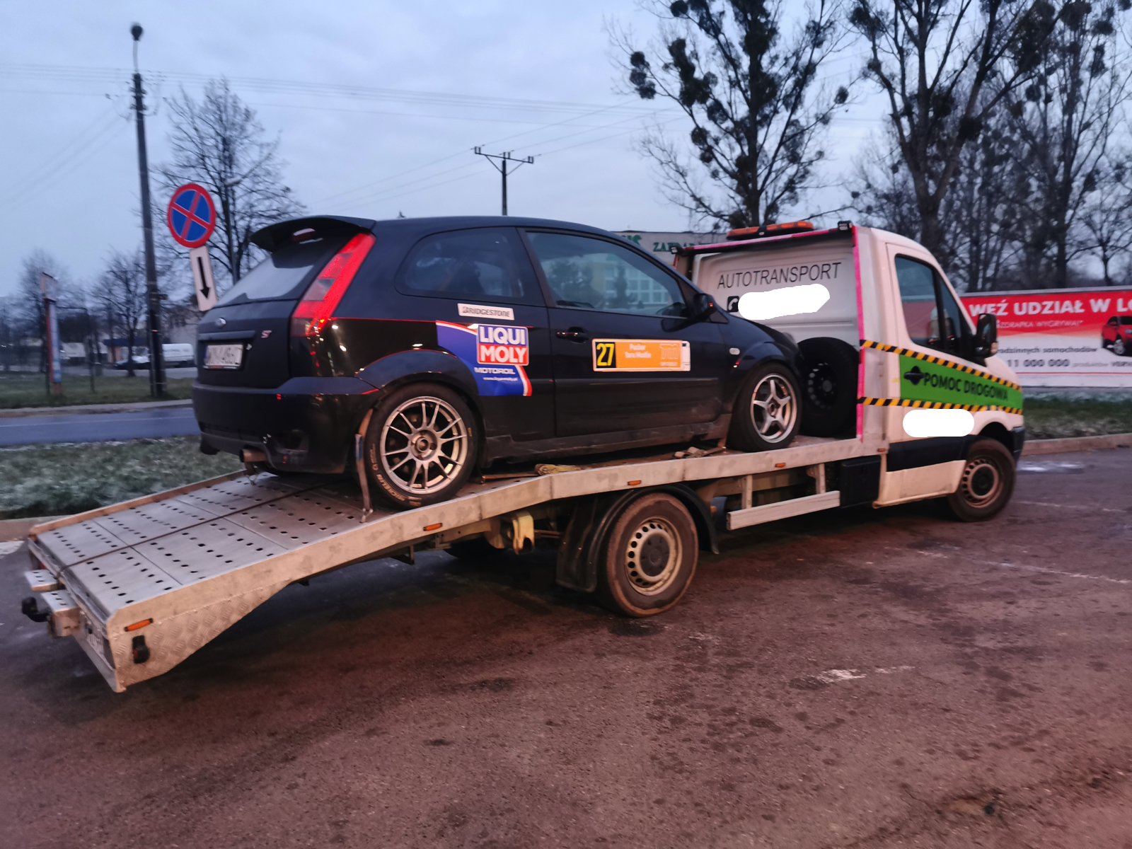 Czarny samochód sportowy z naklejkami sponsorskimi, w tym Liqui Moly i Bridgestone, załadowany na platformę pojazdu pomocy drogowej z napisem 'Autotransport', zaparkowanego na parkingu przy ulicy...