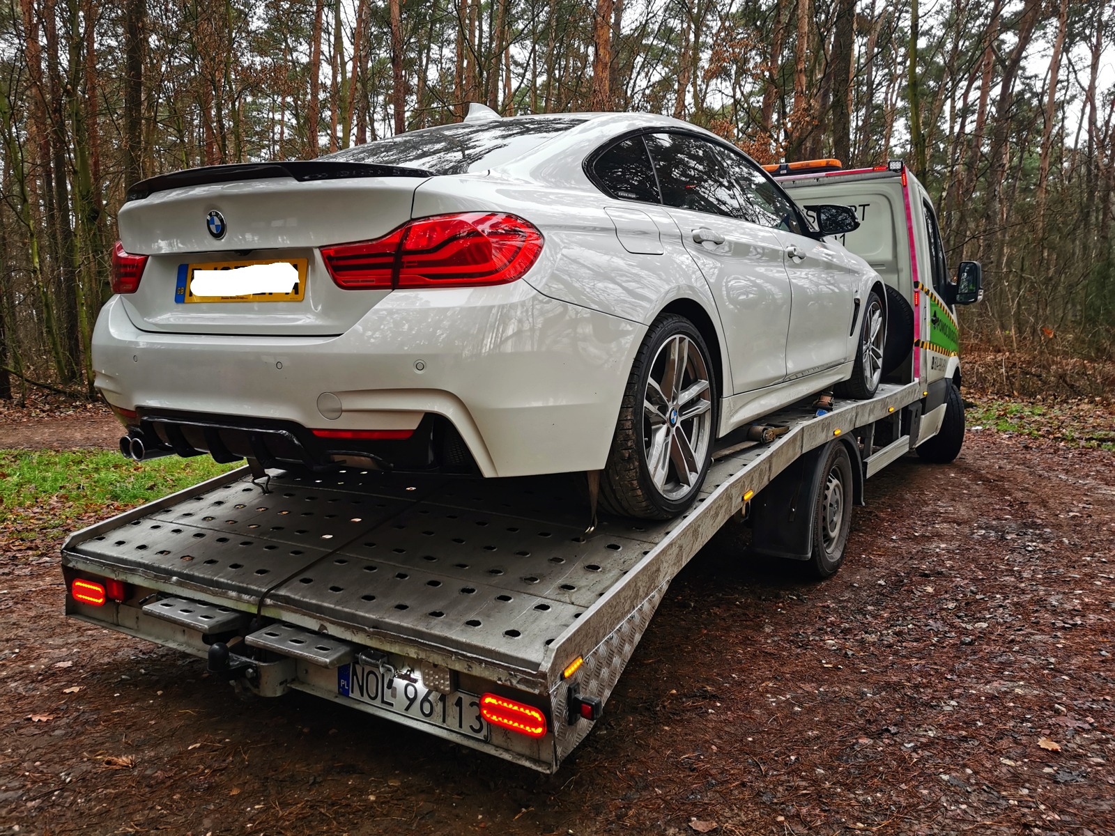 Białe BMW serii 4 na platformie pojazdu pomocy drogowej, widok z tyłu, las w tle.
