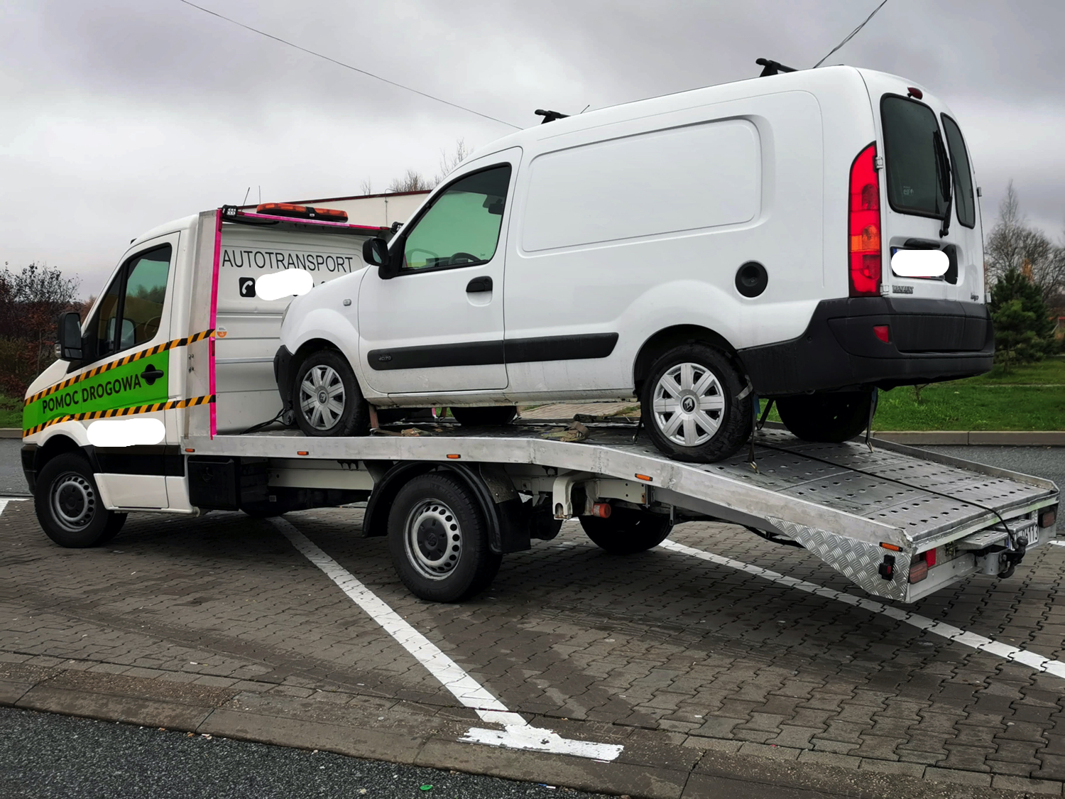 Biały samochód dostawczy transportowany na platformie pojazdu pomocy drogowej z napisem 'Pomoc Drogowa' na boku i 'Autotransport' na kabinie.