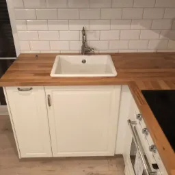 Nowoczesna kuchnia z białymi szafkami, drewnianym blatem, ceramicznym zlewem i białymi płytkami na ścianie; widoczne fragmenty okna i płyty indukcyjnej.