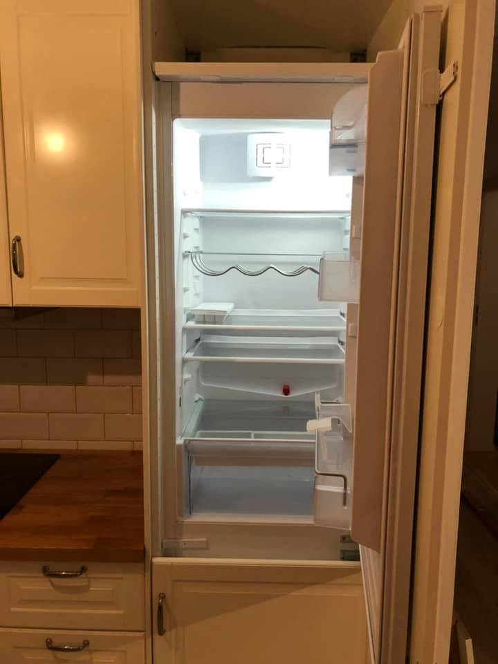 Wbudowana, pusta lodówka z otwartymi drzwiami, zintegrowana z białą zabudową kuchenną i drewnianym blatem, widoczna część ściany z białymi płytkami.