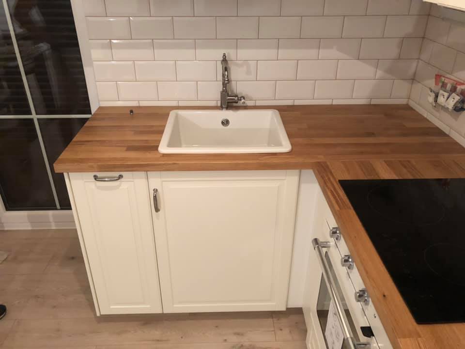 Nowoczesna kuchnia z białymi szafkami, drewnianym blatem, ceramicznym zlewem i białymi płytkami na ścianie; widoczne fragmenty okna i płyty indukcyjnej.