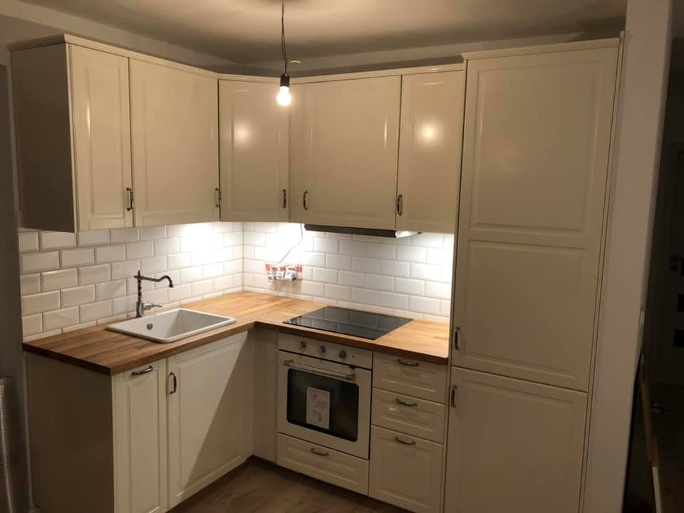 Kompaktowa, jasna kuchnia z białymi szafkami i drewnianym blatem, oświetlona lampką wiszącą nad blatem roboczym i białymi płytkami metra na ścianie.