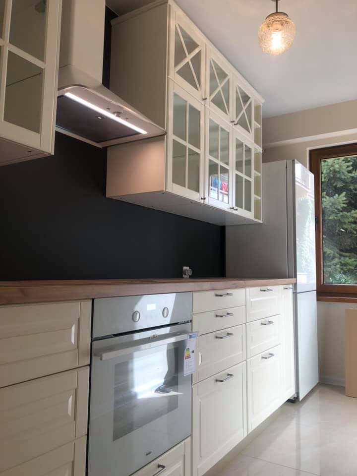 Jasna kuchnia z kremowymi szafkami z przeszklonymi frontami, drewnianym blatem i czarną ścianą, uzupełniona oświetleniem podszafkowym i wiszącą lampą.