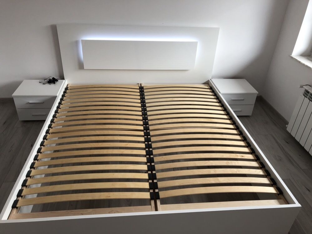 Nowoczesne białe łóżko z drewnianym stelażem i podświetlanym zagłówkiem, z szafkami nocnymi po bokach, ustawione na szarej podłodze w minimalistycznym wnętrzu.