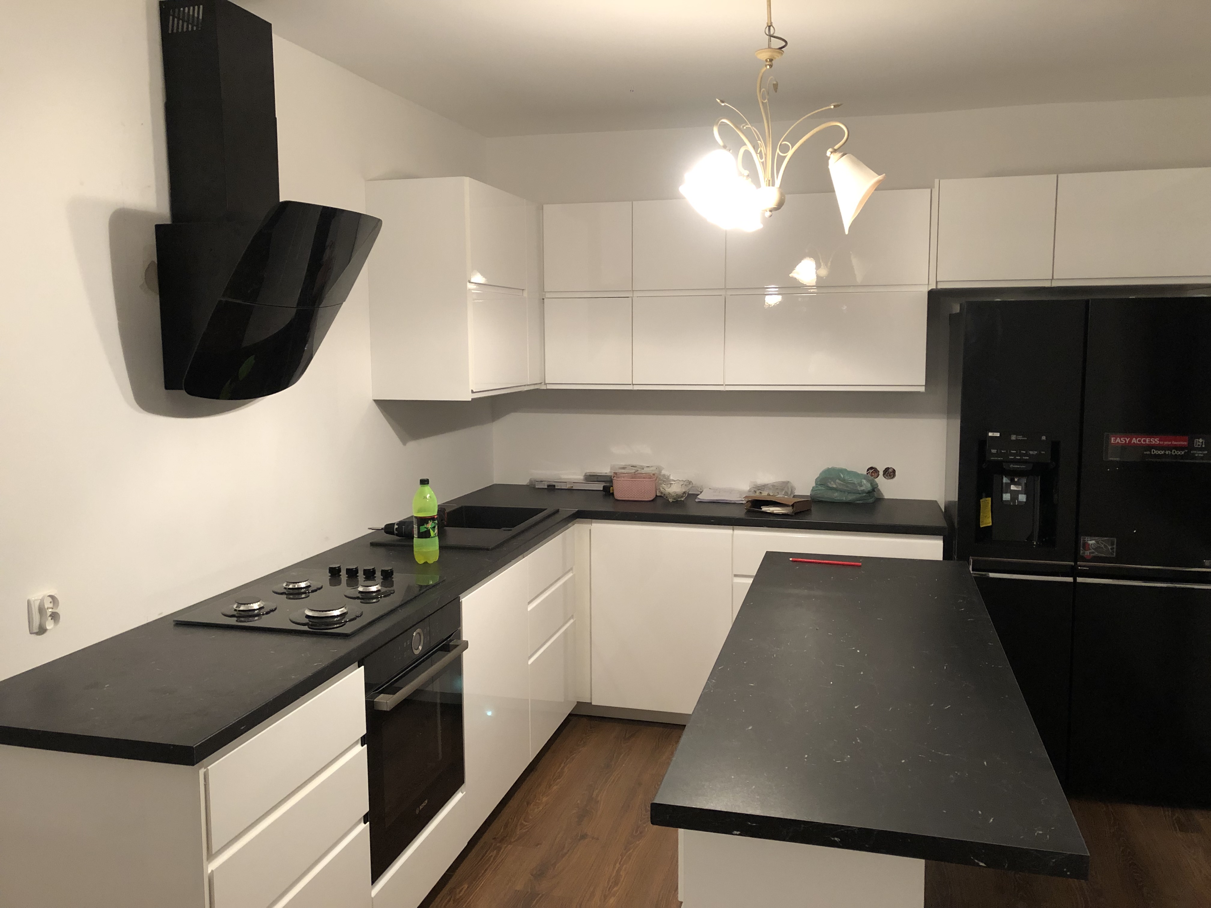 Nowoczesna kuchnia w Nowym Sączu z białymi szafkami, czarnym blatem i sprzętem AGD, w tym piekarnikiem, płytą grzewczą i lodówką side-by-side.